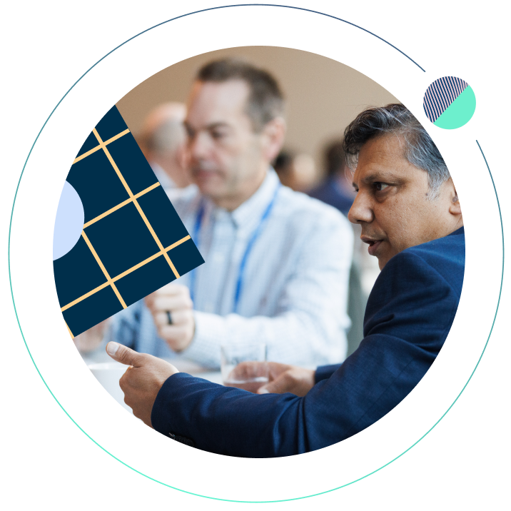 Two conference attendees engaged in conversation at a table, framed in a circular design with a navy grid pattern, yellow diagonal lines, and a small teal accent.
