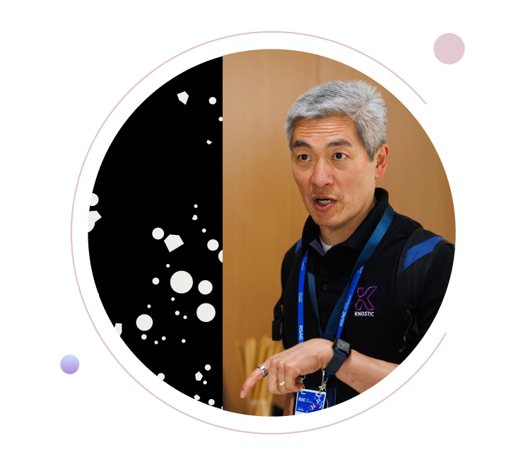 Gray-haired man wearing a black polo shirt and RSAC conference badge, gesturing while speaking, framed by a circular black-and-white geometric design on a purple background.