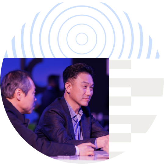 Two men having a discussion at a conference, surrounded by purple geometric shapes and circular patterns.