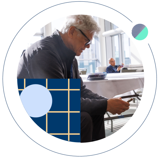 Man with gray hair looking at his phone at a conference, framed by blue geometric patterns.