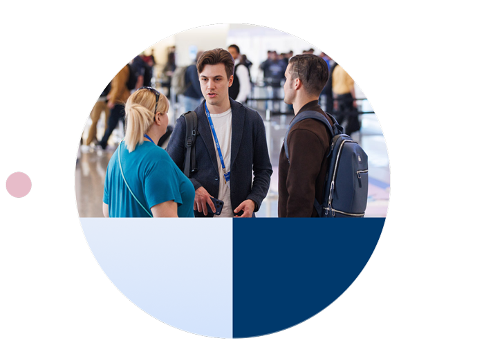 Three conference attendees standing together and having a conversation in a busy convention center, framed by a circular design with light blue and navy geometric sections.
