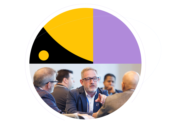 Conference attendees participating in a roundtable discussion, with a man wearing glasses speaking and gesturing, framed by yellow, purple, and black geometric patterns.