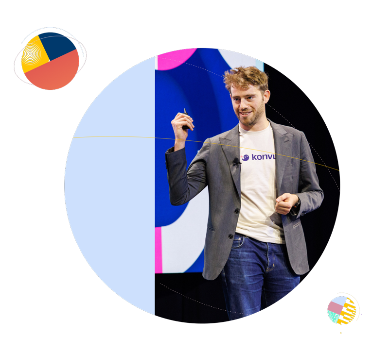 Male speaker wearing a gray blazer and white Konvu T-shirt presenting on stage, framed by light blue and colorful circular geometric designs.