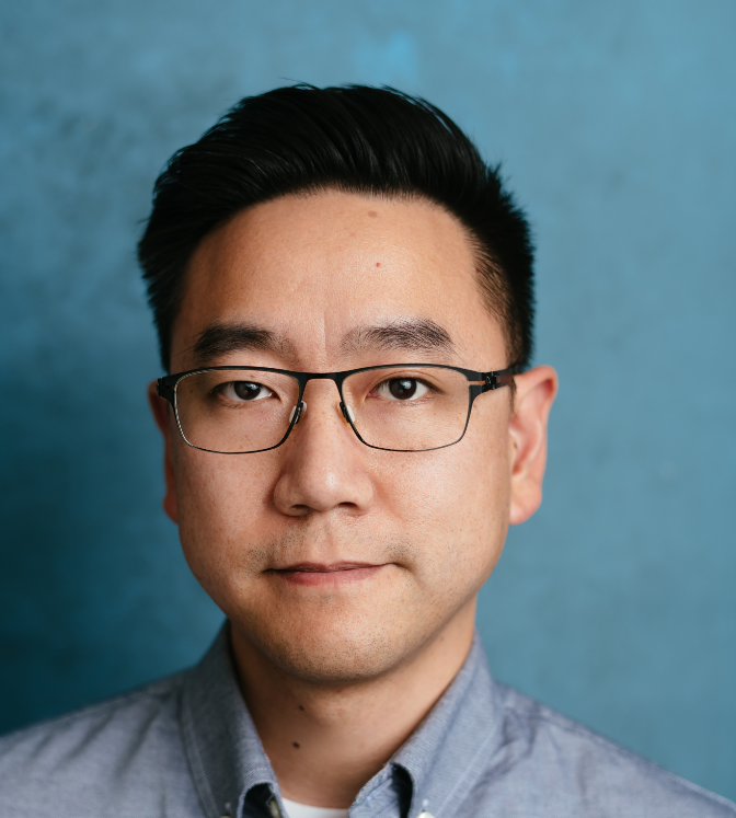 Professional headshot of a man wearing glasses and a gray collared shirt against a blue background.