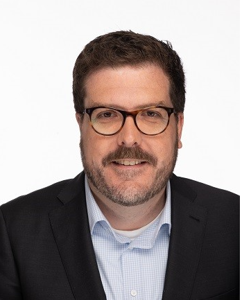 Professional headshot of a man with glasses and a beard wearing a dark blazer.