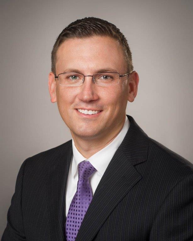 Professional headshot of a smiling man wearing glasses, a dark suit, and a purple tie.