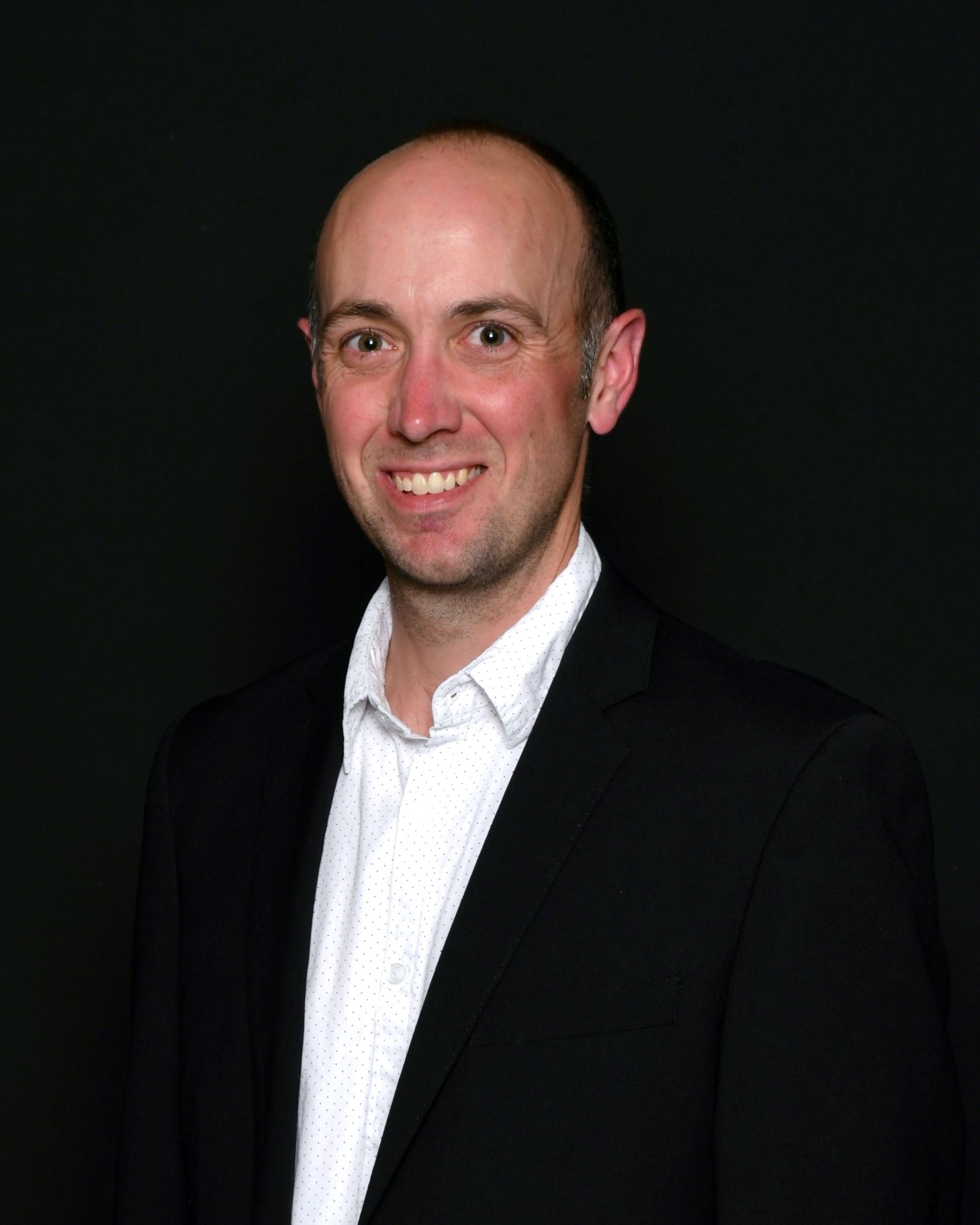 Professional headshot of a smiling bald man wearing a black blazer and white shirt against a dark background.