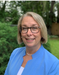 Professional portrait of a smiling woman with blonde hair and glasses wearing a light blue blazer outdoors.
