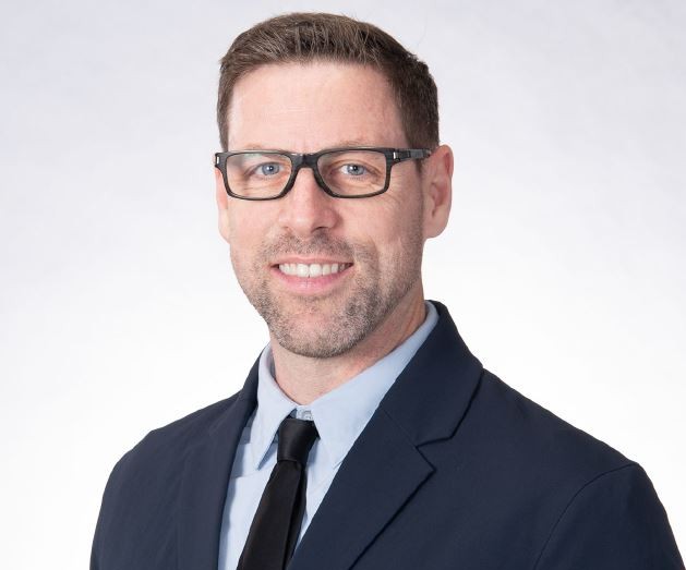 Professional headshot of a man wearing glasses, a navy suit, and a black tie.