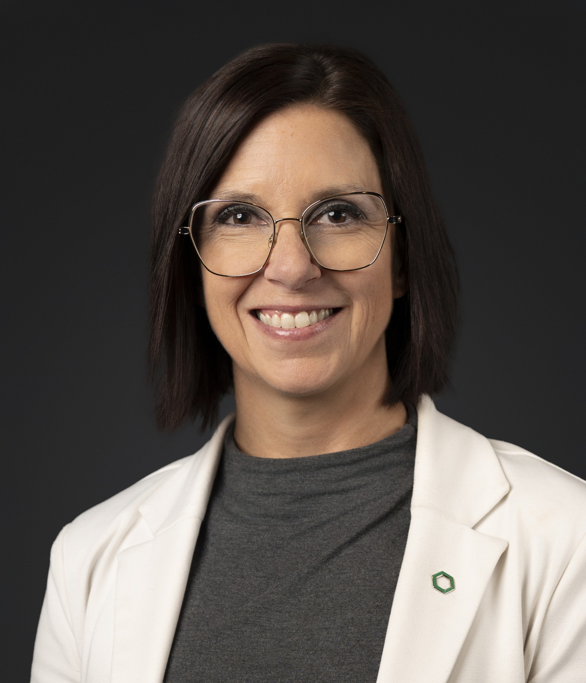 Professional headshot of a smiling woman with dark hair, glasses, and a white blazer.
