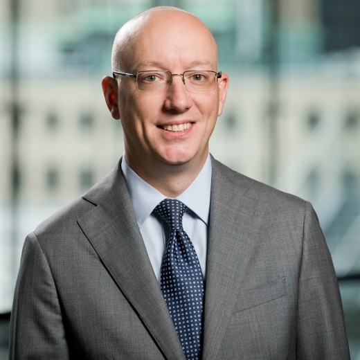 Professional headshot of a smiling bald man wearing glasses, a gray suit, and a blue tie.