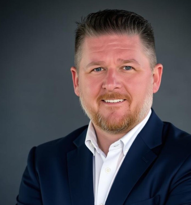 Professional headshot of a man with short hair and a trimmed beard wearing a navy suit.